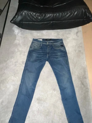 Replay Anbass blå jeans, skinny fit - Säljer ett par Replay Anbass jeans i klassisk blå tvätt med snygga slitningar och skinny passform. Jeansen har fem fickor, Replay-logga på bakfickan och patch i läder bak. Perfekta för dig som gillar en smal siluett och stilren look.