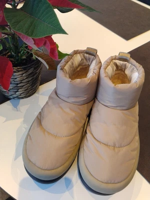 HELT nya Beige vinterboots från UGG 39 - Säljer ett par beige vinterboots från UGG med puffad ovandel och mjukt foder.storlek 39. Skorna har rund tå, platt sula och grovt mönstrad yttersula för extra grepp. Perfekta för kalla dagar och riktigt sköna att ha på sig.