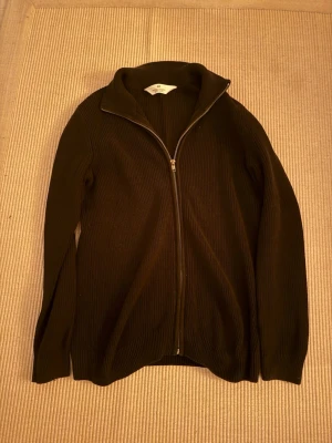 Svart stickad zip från H&M - Säljer en svart ribbad kofta från H&M med hel dragkedja framtill och hög krage. Koftan är långärmad och har en stilren, klassisk look som passar perfekt till lager-på-lager. Materialet är stickat och känns mjukt mot huden.