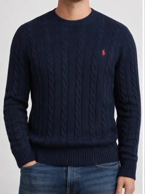 Marinblå kabelstickad tröja Polo Ralph Lauren - Kabelstickad marinblå tröja från Polo Ralph Lauren med rund halsringning och röd broderad logga på bröstet. Tröjan har ribbade muddar och är långärmad, perfekt för kyliga dagar.