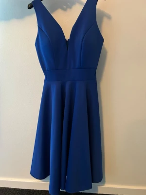 Blå kortklänning med v-ringning - Supersnygg blå kortklänning med djup v-ringning och breda axelband. Klänningen har en markerad midja och ett utsvängt kjolparti som ger en riktigt fin siluett. Tillverkad i ett slätt, lite stretchigt material som känns bekvämt och följsamt.