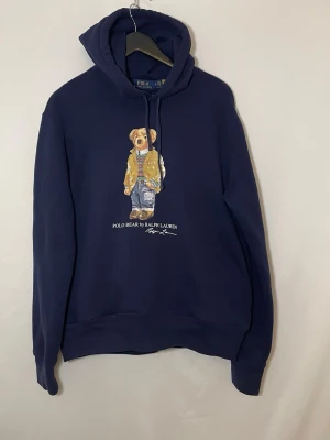 Ralph Lauren hoodie - Mörkblå Ralph Lauren hoodie med klassiskt Polo Bear tryck framtill. Tillverkad i mjuk och bekväm bomullsblandning med huva och känguruficka. Normal passform i storlek M, unisex. Tröjan är i fint skick utan hål eller fläckar. Vid frågor är det bara att höra av😊