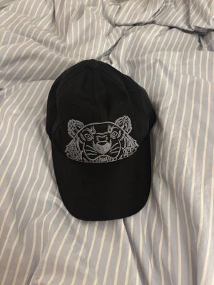 Svart Kenzo keps med tigermotiv - Säljer en svart keps från Kenzo med ikoniskt tigermotiv broderat framtill i grått och Kenzo-logga baktill. Kepsen har klassisk form och justerbar rem. Perfekt accessoar för att lyfta din outfit.