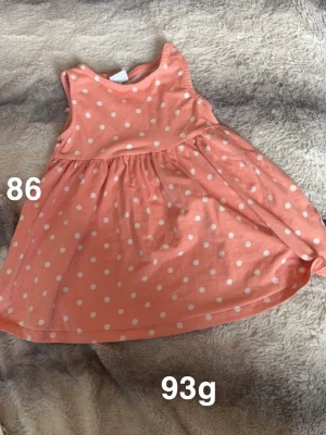 Korallfärgad prickig ärmlös klänning - Supersöt korallfärgad klänning med vita prickar och ärmlös design. Perfekt för varma dagar och enkel att matcha med andra plagg. 🌸 Fint använt skick. Storlek 86