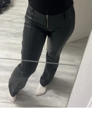 Svarta bootcut byxor i skinnimitation - Snygga svarta byxor i skinnimitation med bootcut-snitt och hög midja. Byxorna har dragkedja framtill, bälteshällor och en stilren look som passar till många olika outfits. Materialet är blankt och ger en cool vibe.