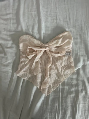 Ljus rosa spets topp  - Säljer en ljusrosa tubtopp i spets med en stor rosett framtill. Toppen är axelbandslös och har ett romantiskt, feminint uttryck med transparent spetsmaterial och knytning. Perfekt för sommarens varma dagar och snygg att styla med jeans eller kjol.