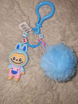 Blå fluffig nyckelring med figur - Söt nyckelring med en blå fluffig boll och en tecknad figur i blå och rosa overall. Nyckelringen har plastdetaljer i blått och pastellfärger samt bokstavspärlor. Perfekt att hänga på väskan eller nycklarna för en lekfull touch.
