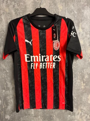 AC Milan  fotbollströja 25/26 - Ny storlek S - AC Milan matchtröja från Puma i storlek S. Tröjan har klassiska röda och svarta ränder med subtilt mönster, broderat klubbmärke och vita Puma-loggor. Tillverkad i lätt polyester som andas, perfekt för fotbollsträning eller att bära på läktaren.