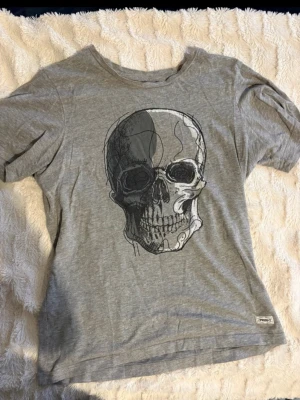 Grå t-shirt med dödskalletryck - Grå t-shirt från en affär utomlands med stort dödskalletryck på framsidan. T-shirten har rund halsringning och korta ärmar. Tillverkad i mjuk bomull och har en avslappnad passform som är perfekt för en cool och edgy stil. Storlek 164.