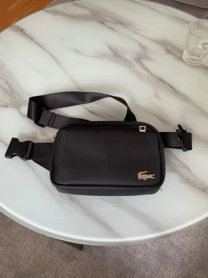 Svart axelväska från Lacoste - Snygg svart axelväska från Lacoste med justerbart band och klassisk krokodil-logga i metall på framsidan. Väskan har en stilren, rektangulär form och stängs med dragkedja. Perfekt för att bära det viktigaste och passar till många olika outfits.