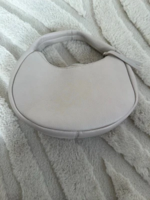 Beige axelväska  - Säljer en stilren beige axelväska, rundad form och dragkedja upptill. Väskan har ett mjukt handtag och minimalistisk design utan synliga loggor eller mönster. Perfekt för dig som gillar en clean och modern look.