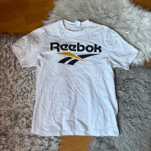 Vit Reebok Classic t-shirt - Vit t-shirt från Reebok Classic med stor svart och gul logga framtill. Klassisk passform med rund hals och korta ärmar. Perfekt till jeans eller shorts för en sportig vibe.