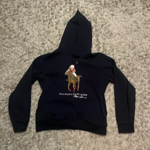 Mörkblå hoodie från Ralph Lauren - Säljer en snygg mörkblå hoodie från Ralph Lauren med ett coolt tryck av en polospelare på framsidan. Perfekt för en avslappnad stil. Hör av dig vid intresse!