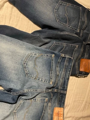 Två par blå jeans från Jack & Jones herr - Säljer två par klassiska blå jeans från Jack & Jones. Ena paret är ljusare med slitningar och tvättade detaljer, medan det andra är mörkare blått. Båda har fem fickor, kontrastsömmar och läderpatch bak i midjan. Perfekta för dig som gillar en avslappnad och stilren look.