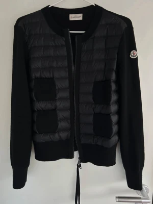 Moncler cardigan  - Svart kofta från Moncler med quiltad front och två fickor. Långärmad med rund hals och dragkedja framtill. Moncler-logga på ärmen. Materialmix med stickade partier och syntet på framsidan. Perfekt för lager-på-lager under kyliga dagar.