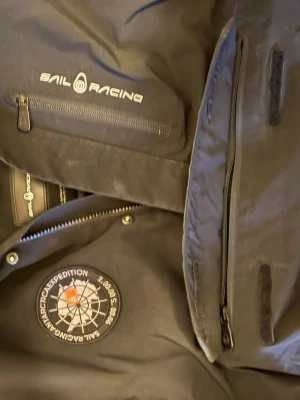 Svart Sail Racing vindjacka med patch - Pris kan diskuteras i dm! Säljer en svart vindjacka från Sail Racing med huva och snygg broderad patch på bröstet. Jackan har dragkedjor, flera fickor. 