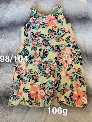 Blommig gul kortklänning - Säljer en söt gul kortklänning med färgglatt blommönster i rosa, grön och svart. Klänningen är ärmlös och har en rund halsringning. Perfekt för varma dagar och ger en riktigt somrig vibe!🌸 Fint använt skick. Storlek 98/104