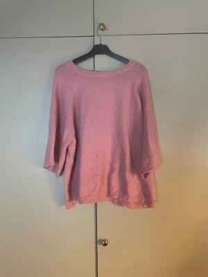 Rosa stickad tröja från 157 - Mysig rosa stickad tröja från 157 med lös passform och trekvartsärm. Tröjan har bred ribbad halsringning och ribbade muddar vid ärmslut och nederkant. Perfekt för lager på lager under kyliga dagar.