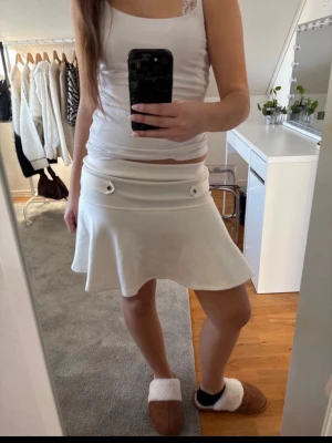 Vit kortkjol från Lindex - Supersnygg vit kortkjol från Lindex med utsvängd form och dekorativa detaljer vid midjan. Kjolen har en clean look och är tillverkad i ett mjukt material som känns bekvämt. Perfekt att styla med crop tops eller stickade tröjor.