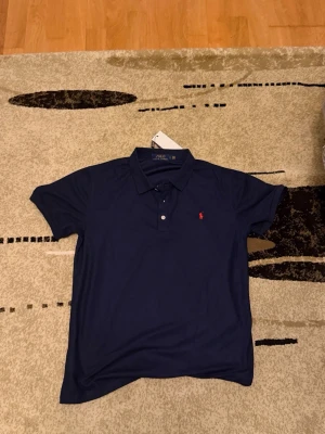 Marinblå pikétröja från Ralph Lauren - Säljer en klassisk marinblå pikétröja från Polo Ralph Lauren. Tröjan har korta ärmar, krage och två knappar framtill. På bröstet finns den ikoniska röda Polo-loggan broderad. Materialet är mjukt och andas, perfekt för en clean och stilren look.