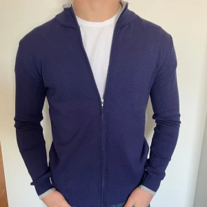 Marinblå kofta med dragkedja - Snygg marinblå kofta med dragkedja framtill. Tröjan är långärmad och har ribbade muddar i ljusgrått vid ärmslut och hals. Perfekt att slänga på sig över en t-shirt för en clean och enkel look.