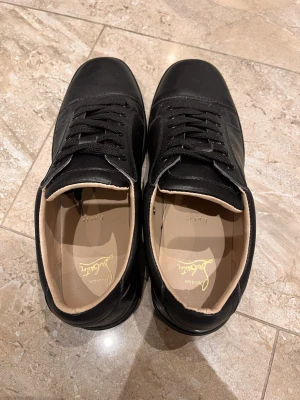 Svarta sneakers från Christian Louboutin - Säljer ett par svarta sneakers från Christian Louboutin i skinn med klassiska röda sulor. Skorna har rund tå, svarta skosnören och beige innersida. Kommer med originalpåse. Perfekta för dig som vill ha en clean och lyxig look.