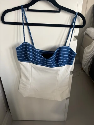 Randig blå och vit linnetopp XS - Snygg linnetopp med smala axelband och bystparti i marinblått med vita ränder. Nedre delen är helvit och toppen har en figurnära passform. Perfekt för sommardagar och enkel att matcha med jeans eller kjol.