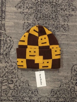 Brun och gul rutig mössa Acne Studios - Stickad mössa från Acne Studios i brunt och gult med rutigt mönster och smiley-ansikten. Mössan är i 100% ull och har en vikt kant med Acne Studios logopatch framtill. Perfekt accessoar för att sticka ut i mängden.