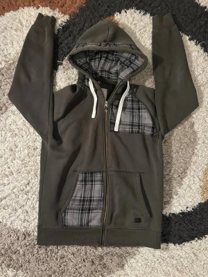 Grön zip up  - Grön zip up hoodie i storlek M, men passar mer som S. Vanlig hoodie som jag har sytt om med symaskin och lagt till extra material i rutigt tyg på ficka, bröst och insida av huvan. Unik och bekväm, bra skick.