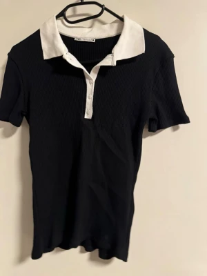 Svart ribbad polotopp från Zara - Säljer en svart ribbad polotopp från Zara med vit krage och knappslå. Toppen har korta ärmar och en klassisk piké-look med kontrastfärgade detaljer. Perfekt för en stilren och sportig vibe.