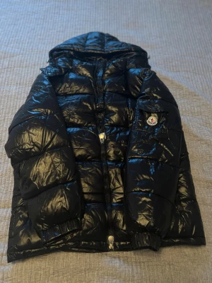 Svart Moncler pufferjacka med huva - Säljer en svart, glansig pufferjacka från Moncler med huva och dragkedja framtill. Jackan har en ficka med Moncler-logga på ärmen och quiltad design. Perfekt för kalla dagar och riktigt snygg till streetwear-stilen. NFC funkar samt QR koden