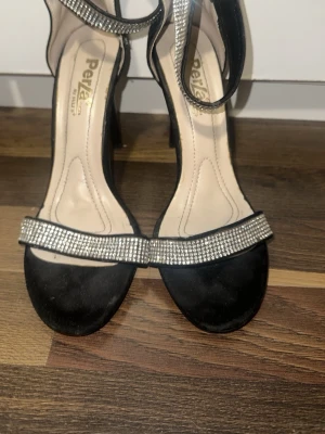 Svarta sandaler med strass och klack - Snygga svarta sandaler från Perla by Call It Spring med hög klack och öppen tå. Remmarna är dekorerade med glittrande strass och skon har dragkedja bak för enkel påtagning. Materialet är mocka och syntet, perfekt för dig som vill sticka ut.