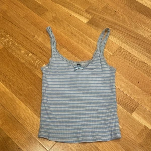 Blåvit randig spetslinnetopp - Supersöt blåvit randig linnetopp från H&M med spetskant och liten rosett framtill. Toppen är ribbad och har smala axelband med spetsdetaljer. Perfekt för sommardagar.