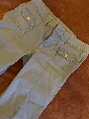Ljusbeiga bootcut cargopants - Säljer ett par ljusblå cargopants med bootcut passform. Byxorna har två fickor framtill med lock och guldknappar, breda hällor och raka sömmar längs benen. Materialet känns mjukt och lätt, perfekt för en chill och avslappnad stil.