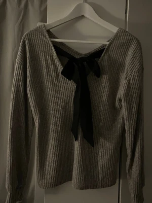 Grå ribbad tröja med rosett - Säljer en grå ribbad tröja med lång ärm och snygg v-ringning i ryggen. Tröjan har en svart rosett som detalj i nacken och är gjord i ett mjukt stickat material. Perfekt för dig som gillar enkla men ändå unika plagg.