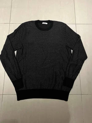 Svart stickad tröja från J.Lindeberg - Säljer en svart långärmad tröja från J.Lindeberg i storlek M. Tröjan har ett diskret rutigt mönster i grått och svarta ribbade muddar vid ärmslut, hals och soppslut. Perfekt till höst och vinter för en clean och stilren look. Nopprig men går att lösa!!