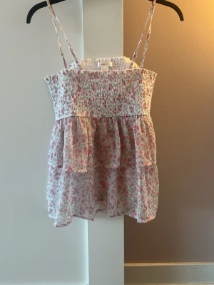 Blommig volangtop från Gina Tricot - Supersöt blommig topp från Gina Tricot med tunna axelband och smockad överdel. Toppen har volanger och är gjord i ett lätt, luftigt tyg i vitt med rosa och gröna blommor. Perfekt för varma dagar och ger en romantisk vibe.denna är aldrig använd,super fin nu till våren och sommaren