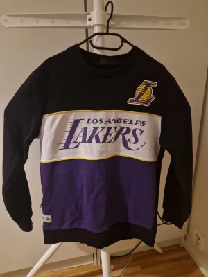 Los Angeles Lakers NBA tröja L - Snygg Los Angeles Lakers crewneck i svart, vitt och lila med stort Lakers-tryck och logga på bröstet. Tröjan har ribbade muddar och är tillverkad i mjukt bomullsmaterial. Perfekt för dig som älskar basket och vill ha en sportig stil.