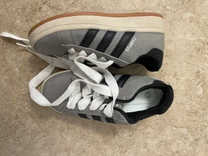 Adidas Campus grå sneakers strl 37 - Säljer ett par Adidas Campus sneakers i grå mocka med svarta läderdetaljer och klassiska tre ränder på sidan. Skorna har vit sula, bruna yttersulor och vita snören. Perfekta för dig som gillar en clean och tidlös stil.