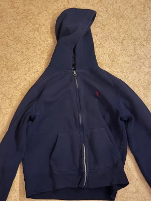 Mörkblå hoodie från Polo Ralph Lauren - Mörkblå hoodie med dragkedja från Polo Ralph Lauren. Klassisk design med huva, känguruficka och röd broderad logga på bröstet. Mjuk insida i bomullsblandning, perfekt för chill dagar. Snygg och enkel att matcha med jeans eller joggers.