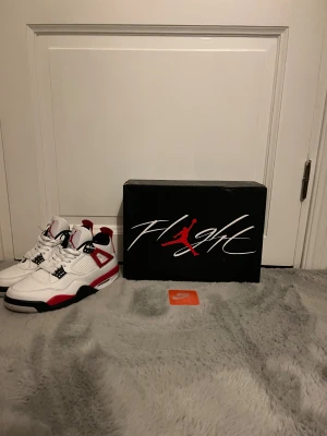 Nike Air Jordan 4 red cement - Nike Air Jordan 4 red cement sneakers med svarta och röda detaljer och den klassiska Jordan-loggan på plösen och yttersulan. Tillverkade i skinn och syntetmaterial, med platt sula och rund tå.