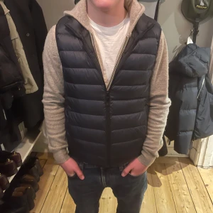 Mörkblå pufferväst från Uniqlo - Snygg svart pufferväst med horisontella quiltade paneler och dragkedja framtill från Uniqlo. Västen har en clean look och passar perfekt över en hoodie eller tröja. Materialet är syntet och designen är normal passform, vilket gör den enkel att styla till jeans eller sweatpants.