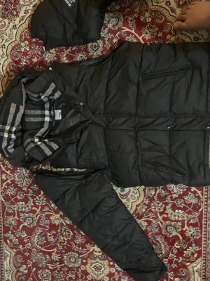 Svart pufferjacka från Burberry - Svart pufferjacka från Burberry med avtagbar huva och klassiskt rutigt foder i svart, vitt och rött. Jackan har dragkedja, tryckknappar och flera fickor med dragkedja. Perfekt för kalla dagar och har en riktigt snygg streetstil.