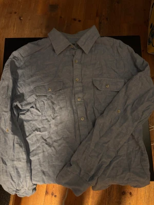 Blå linneskjorta Dressmann L - Snygg blå skjorta från Dressmann i linnemix, storlek L. Klassisk krage, två bröstfickor med knappar och långa ärmar. Perfekt för dig som gillar en chill och stilren look. Skjortan har en lätt och luftig känsla tack vare materialet.