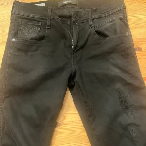 Svarta Replay Anbass jeans - Säljer ett par svarta Replay Anbass jeans med slim fit och snygg slitning på ena knät. Klassisk femficksmodell med dragkedja och knapp framtill. Jeansen är i stretchigt material som sitter tajt och bekvämt. Perfekta för dig som gillar en modern och stilren look. Storlek 29/32!