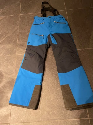 Blå täckbyxor från 8848 Altitude - Snygga blå och svarta täckbyxor från 8848 Altitude med justerbara hängslen, flera dragkedjefickor och förstärkta benslut. Perfekta för vintern och outdoor-aktiviteter. Funktionell design med coola detaljer och logga på benet.