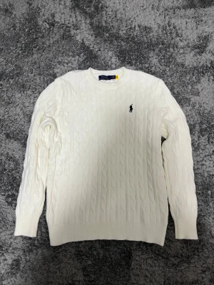 Vit kabelstickad tröja Polo Ralph Lauren - Vit kabelstickad tröja från Polo Ralph Lauren med klassisk rund hals och svart broderad logga på bröstet. Tröjan har långa ärmar och ribbade muddar vid ärmslut och nederkant. Perfekt för dig som gillar stilren och tidlös design.