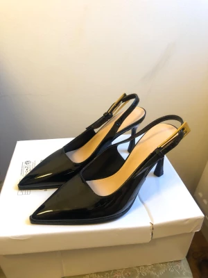 Svarta lackpumps med spetsig tå - Snygga svarta pumps i lack med spetsig tå och hög klack. Skorna har en öppen häl med justerbart spänne i guld och en elegant, smal siluett. Perfekta för dig som gillar stilrena och trendiga skor med lite edge.