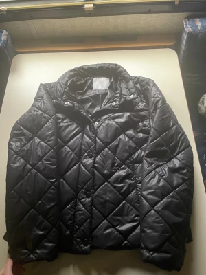 Svart quiltad pufferjacka Samsa S - Quiltad svart pufferjacka från Samsa Collection i storlek S. Jackan har ett snyggt rutmönster,krage och knäppning framtill. Materialet är lite glansigt och känns som syntet, perfekt för kyliga dagar. Stilren och enkel att matcha med din garderob.