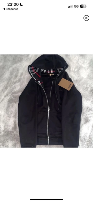 Svart hoodie från Burberry med dragkedja - Svart hoodie från Burberry med klassiskt rutigt foder i huvan i svart, vitt och rött. Tröjan har snörning vid huvan, två fickor fram och hel dragkedja. Perfekt för dig som gillar stilrena och lyxiga streetwear-plagg.